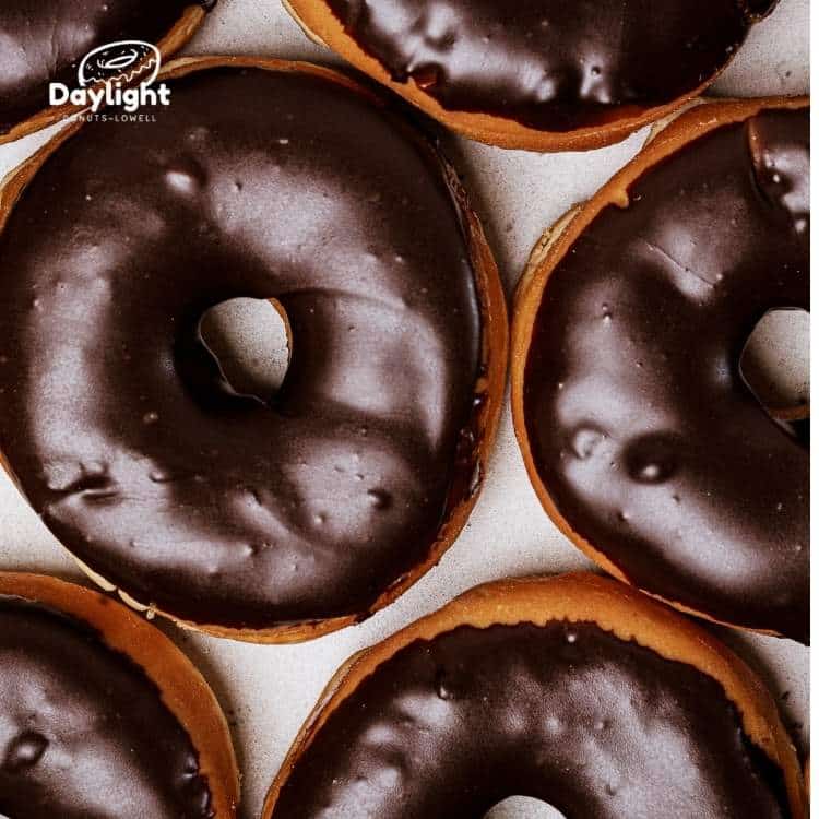 A dozen chocolate glazed donuts from Daylight Donuts Lowell, soft and golden, covered with a rich chocolate glaze.