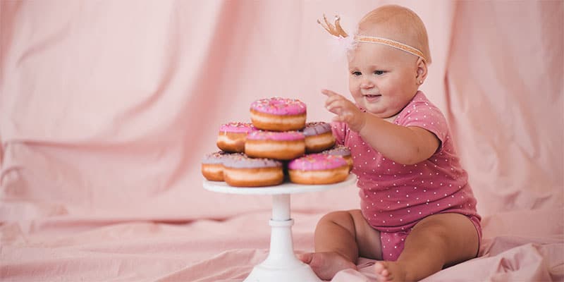 Baby girl celebrating her donut-themed first birthday, sitting next to a cake stand with pink frosted donuts, capturing a perfect moment.