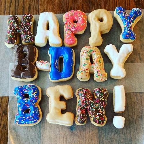 Happy B-Day Ben spelled out in colorful frosted donuts with sprinkles on a wooden surface.