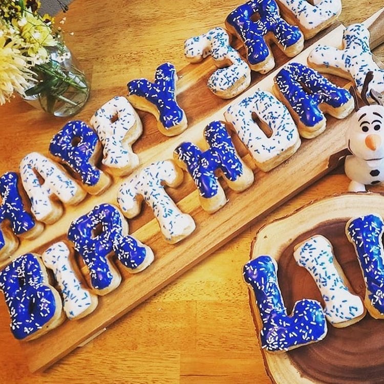 Happy 3rd birthday spelled out in blue and white frosted donuts, with a Frozen-themed Olaf figurine on a wooden table.